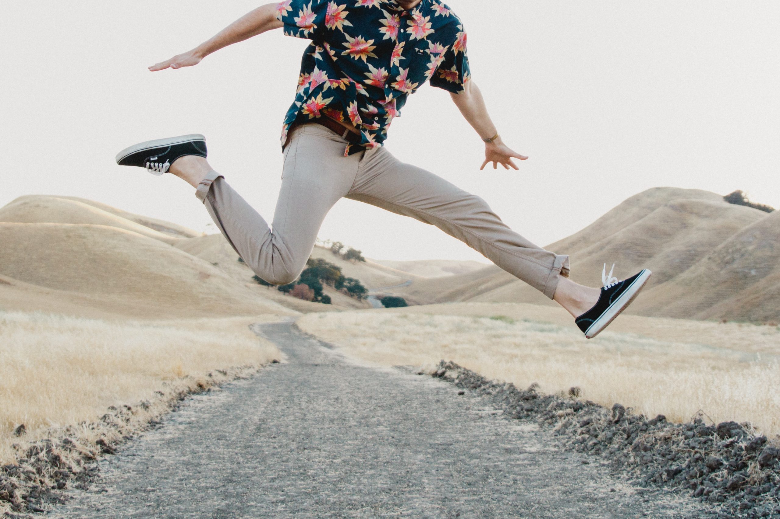 Man in floral shirt leaping over a path on a hilly desolate area.