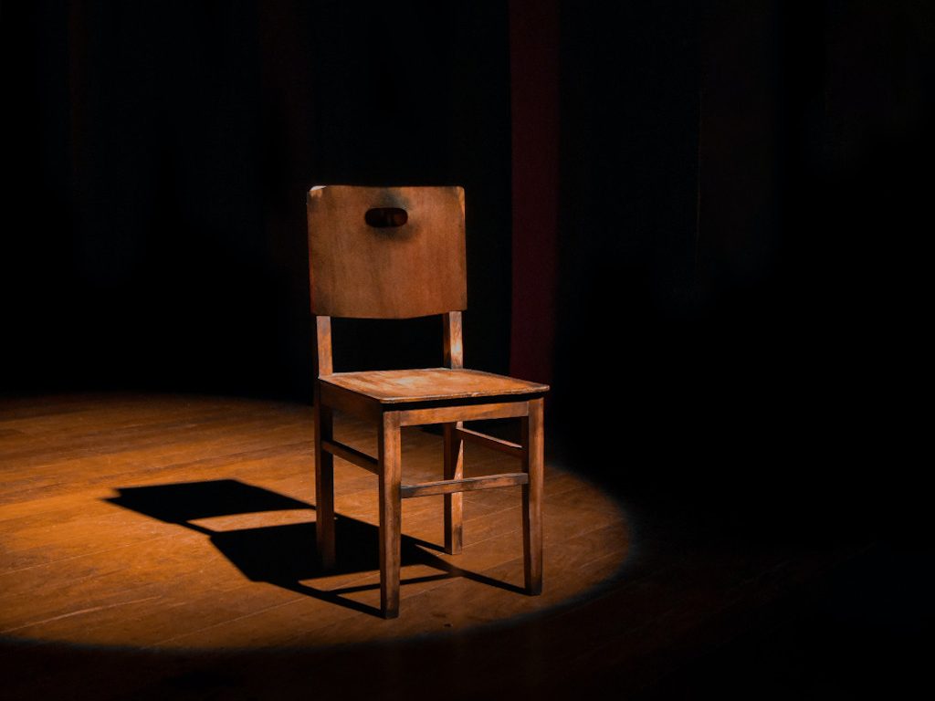 4-legged wooden chair, illuminated under a spotlight on a darkened stage
