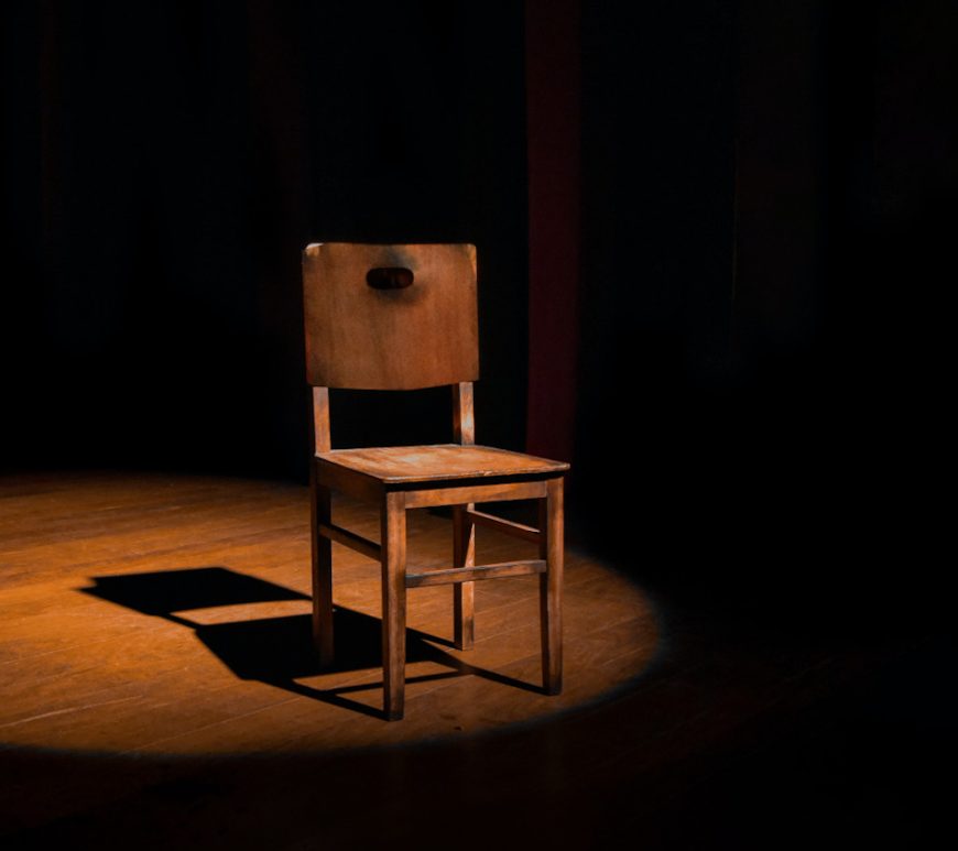 4-legged wooden chair, illuminated under a spotlight on a darkened stage