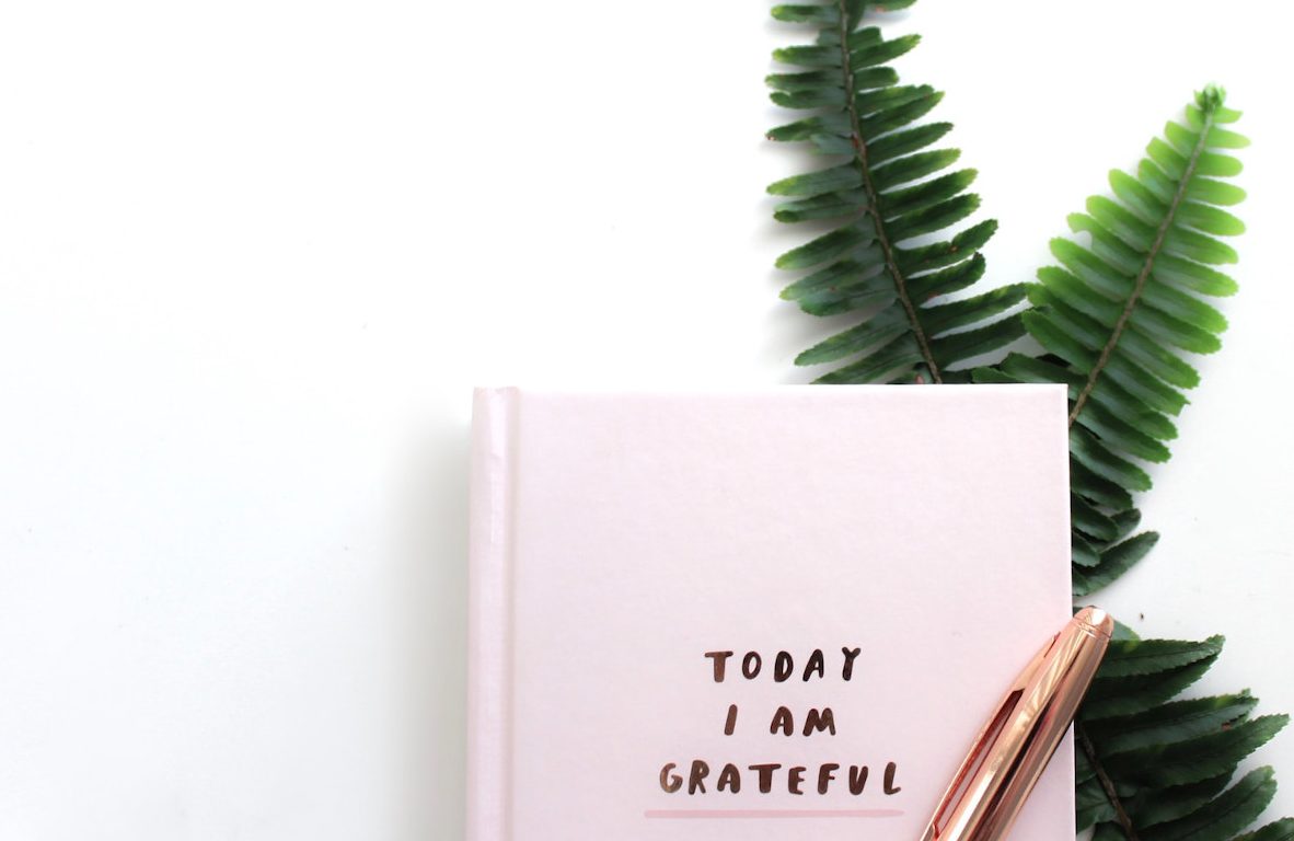 Pink journal with title "Today I Am Grateful" with a rose-gold pen on top and ferns beneath.