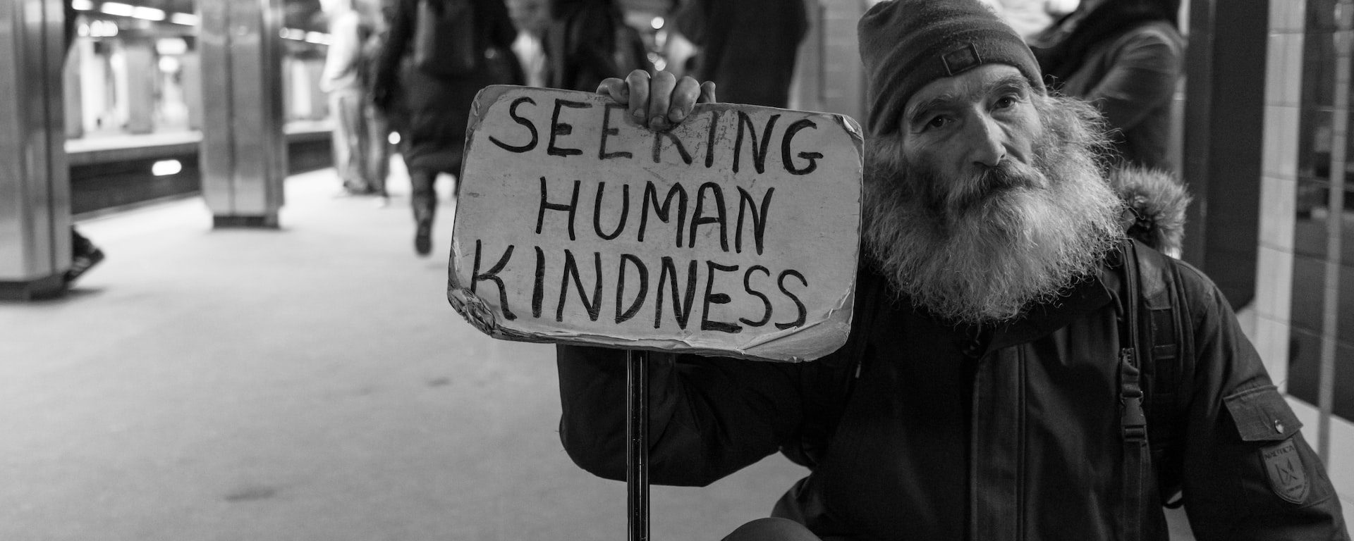 Black and white image of an older man squatting on metro platform. He has a full, grey beard and is holding a sign stating "Seeking Human Kindness". People waiting for a train in the background.