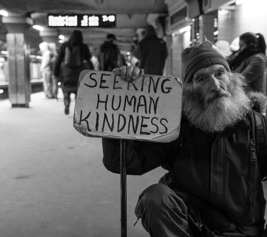 Black and white image of an older man squatting on metro platform. He has a full, grey beard and is holding a sign stating "Seeking Human Kindness". People waiting for a train in the background.