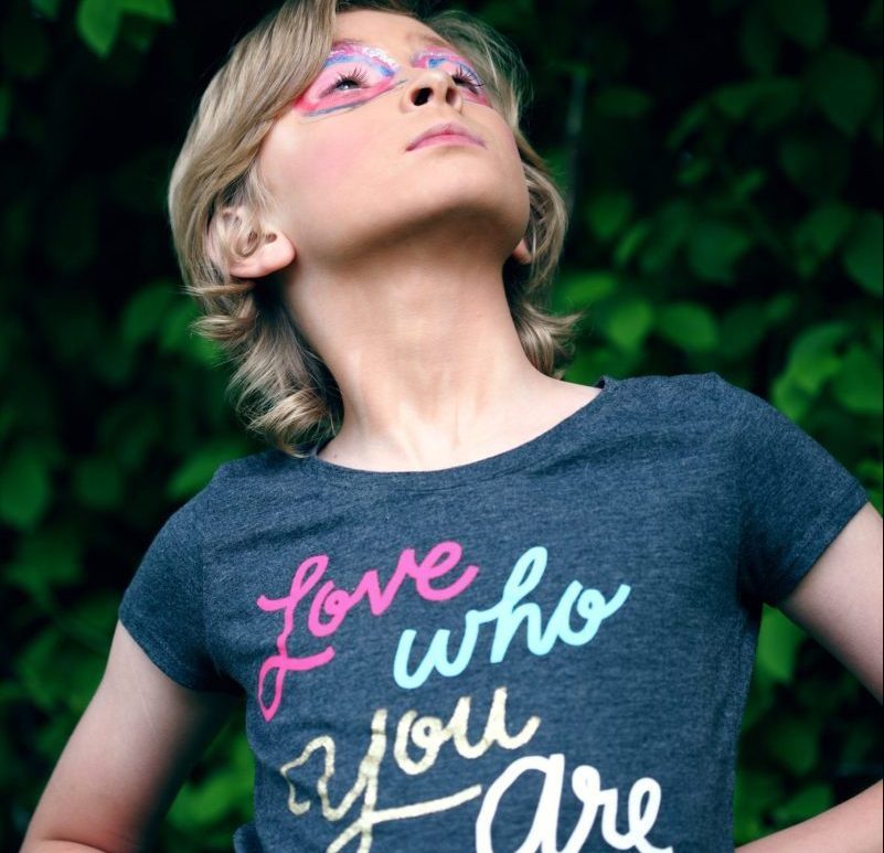 Female standing with hands on waist looking upwards. Make-up and glitter create goggles around her eyes. She is wearing a t-shirt with the expression "Love who you are".
