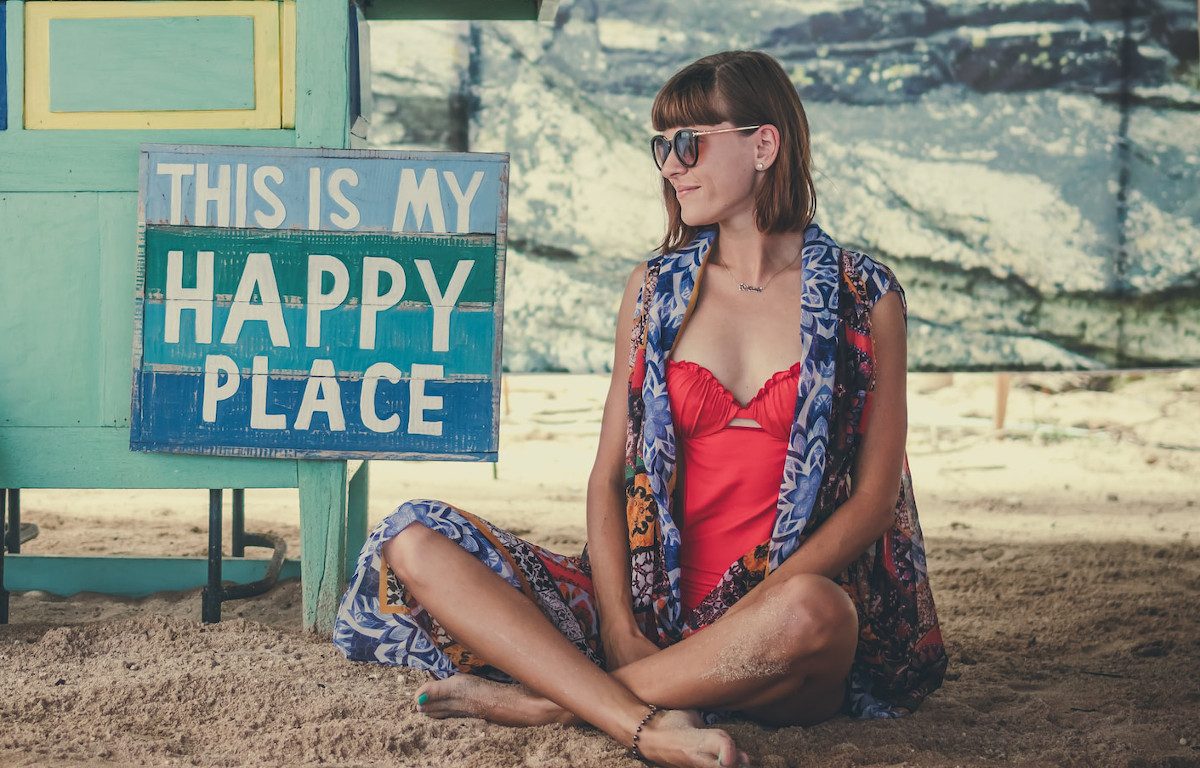 Smiling woman in red bathing suit, patterned blue cover up, and sunglasses, sitting cross-legged in the sand. Striped blue wood sign next to her says "This is my Happy Place" in white font.