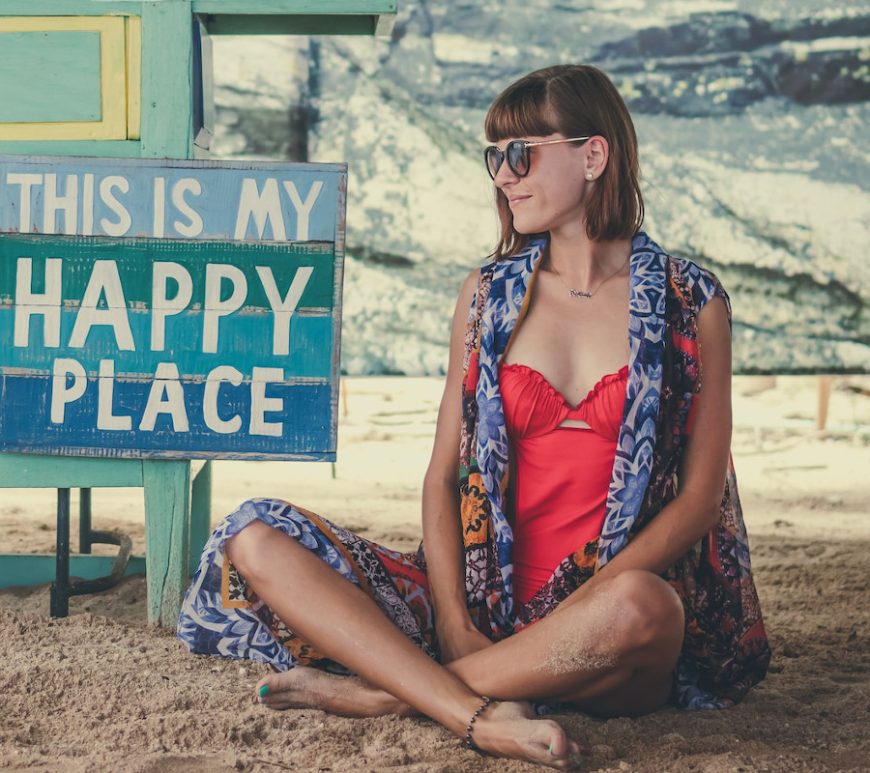 Smiling woman in red bathing suit, patterned blue cover up, and sunglasses, sitting cross-legged in the sand. Striped blue wood sign next to her says "This is my Happy Place" in white font.