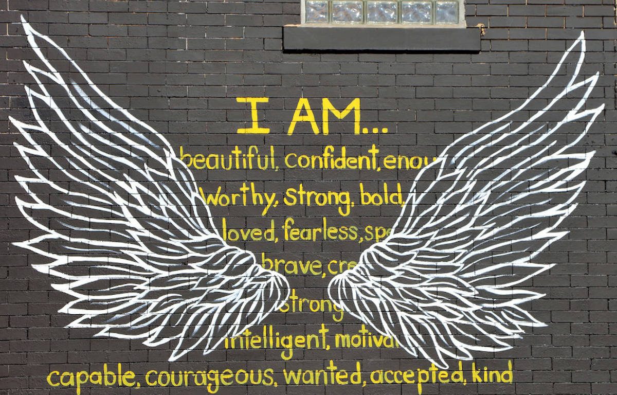 Gray brick wall with graffitti white wings overtop of yellow font affirmation statement "I AM...beautiful, confident, enough, worthy, strong, bold, loved, fearless, special, brave, creative, strong, intelligent, motivated, capable, courageous, wanted, accepted, kind".