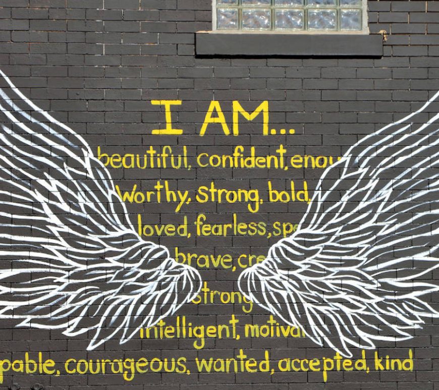 Gray brick wall with graffitti white wings overtop of yellow font affirmation statement "I AM...beautiful, confident, enough, worthy, strong, bold, loved, fearless, special, brave, creative, strong, intelligent, motivated, capable, courageous, wanted, accepted, kind".