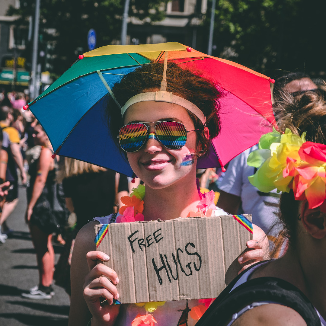 In an outdoor crowd, young smiling adult holds a cardboard sign. They are wearing aviator rainbow sunglasses, rainbow lei, and rainbow umbrella hat. The sign says "Free Hugs".