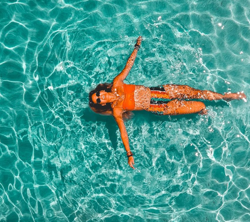 Woman floating on clear, teal water wearing orange top and stripped, multi-colour bottom.