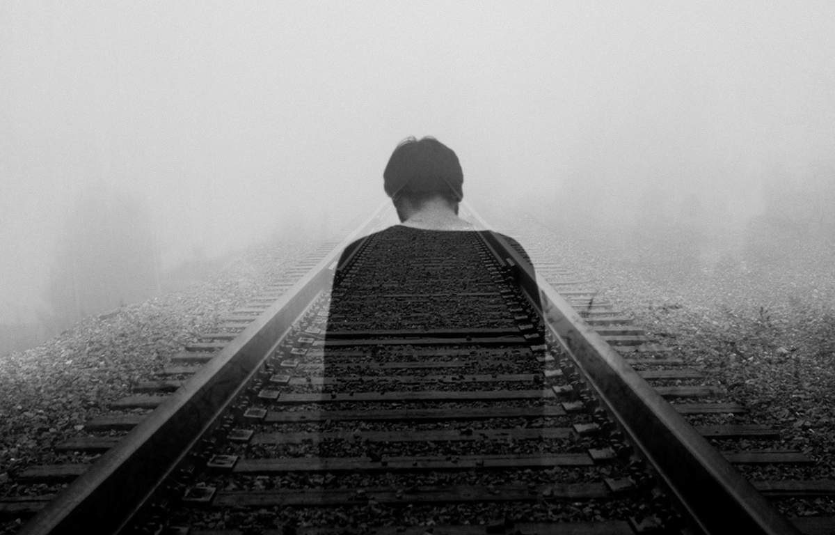 Overexposed black and white image of sad man standing on train tracks. It is foggy, trees are faintly visible in the background.