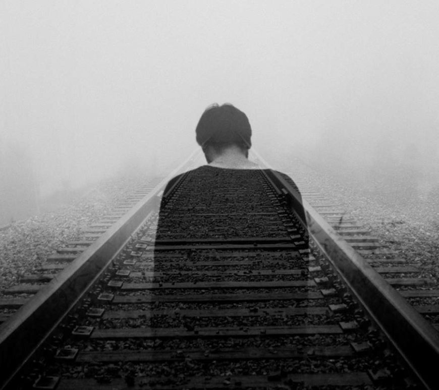 Overexposed black and white image of sad man standing on train tracks. It is foggy, trees are faintly visible in the background.
