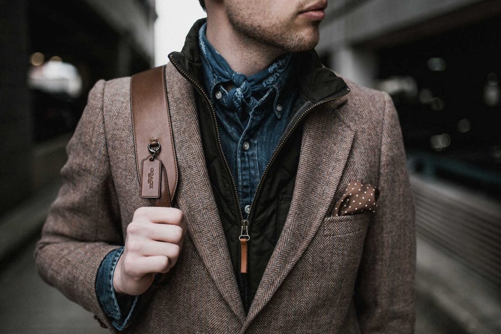 Man walking by shops looking at the windows. He has a denim shirt on, a scarf on his neck, a dark brown vest with a leather zipper-pull, a tweed brown blazer, a pocket square in brown and white polka dot, and a leather Coach bag over his right shoulder.