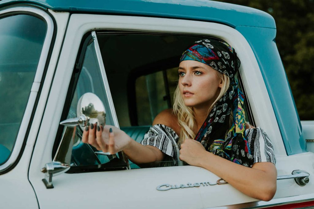 Young blonde woman looking at herself in the side mirror of her turquoise pickup. She is wearing a black-and-white stripped, off-the-shoulder top with a multi-coloured scarf on her head.