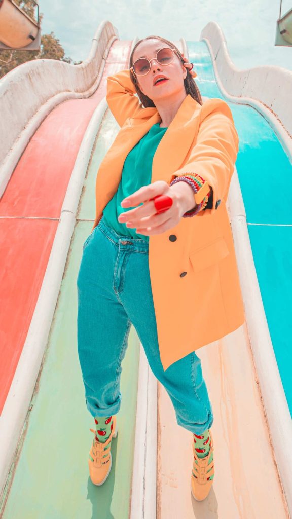 Woman standing on giant slide wearing sunglasses. Colours of the slide and her outfit are various tones of orange and teal.