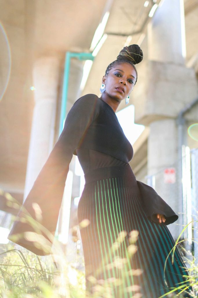 Black woman with bun on her head. She is standing under a bridge, the sun shines behind her. She is wearing a black, bell-sleeve top, earrings, and a black and green striped skirt.