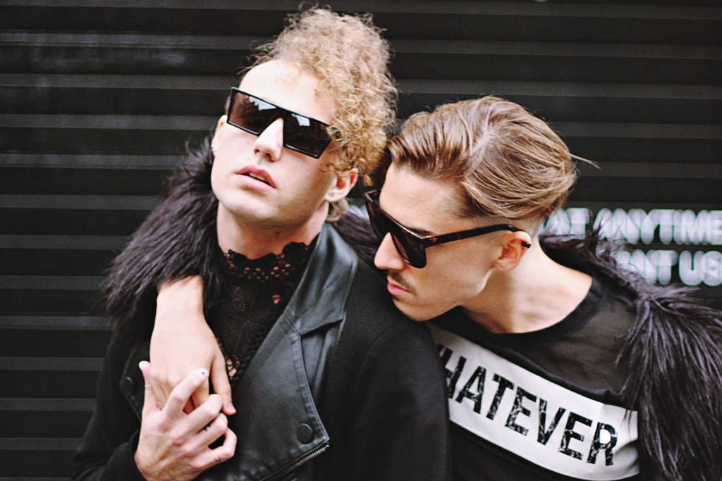 Two men in black holding hands wearing sunglasses. One has a leather coat, the other a fur coat with a T-shirt that says "Whatever".