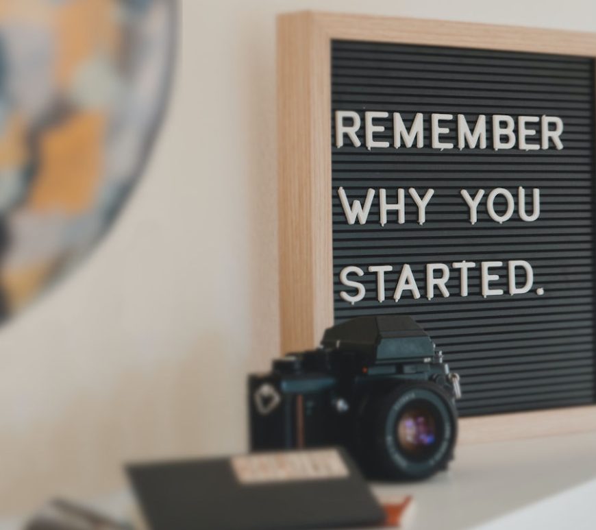 Photo of shelf has a notebook, a camera, and a sign that reads "Remember why you started.".