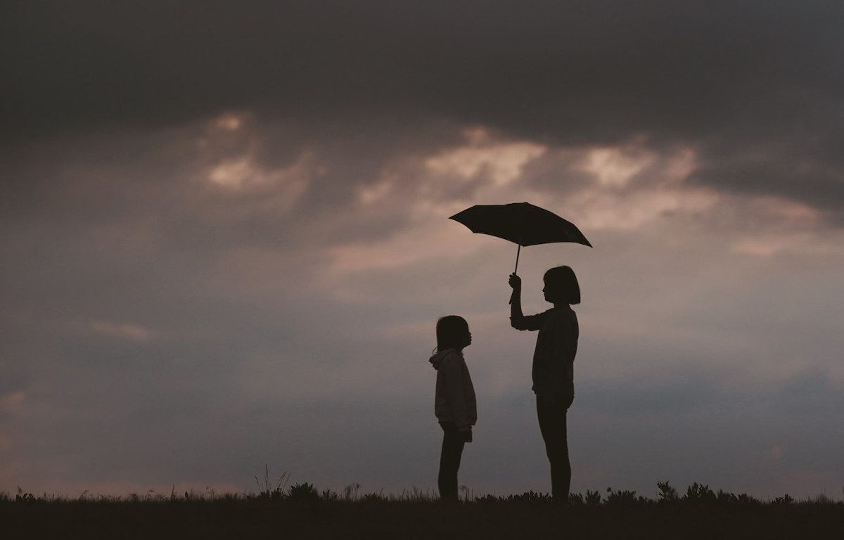 Silhouette of an adolescent girl holding an umbrella over a younger girl. They are in a grass field under cloudy, stormy skies.