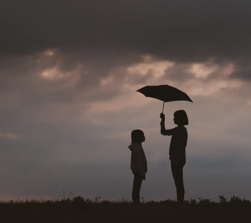 Silhouette of an adolescent girl holding an umbrella over a younger girl. They are in a grass field under cloudy, stormy skies.