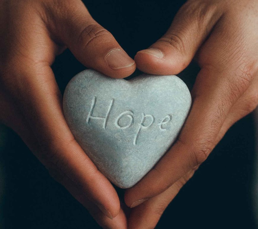 Pair of hands holding a heart-shaped rock with the word hope engraved on the surface.