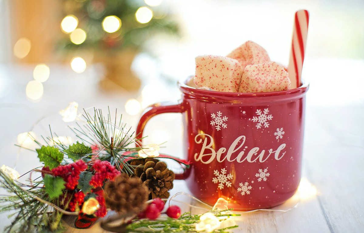 Red mug on a table next to pine cones, red berries, and white holiday lights. White font on the mug reads "Believe", accompanied by snowflakes. Peppermint marshmallows and a candy cane stick out from the top.