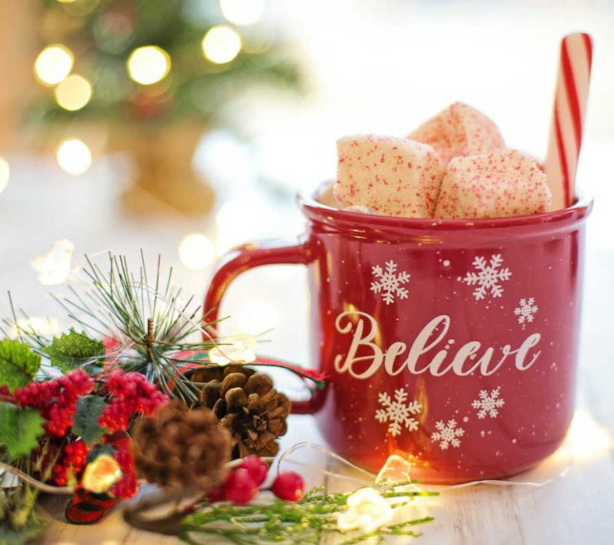 Red mug on a table next to pine cones, red berries, and white holiday lights. White font on the mug reads "Believe", accompanied by snowflakes. Peppermint marshmallows and a candy cane stick out from the top.