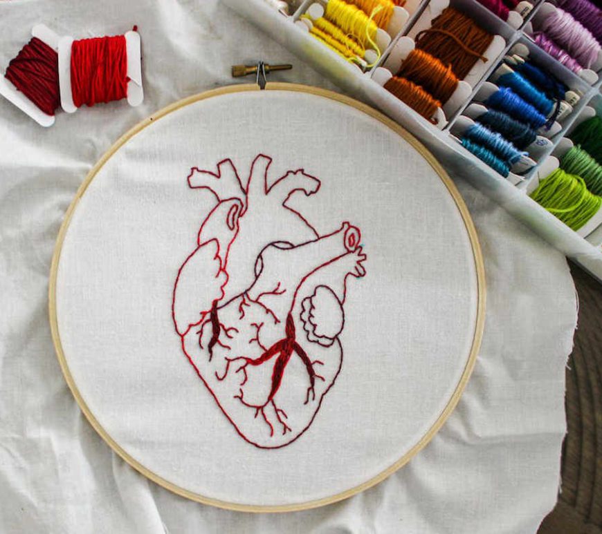 Round embroidery hoop with white cloth and crimson and red embroidered heart. In the upper right corner, a plastic organizer box with embroidery floss in assorted colours.