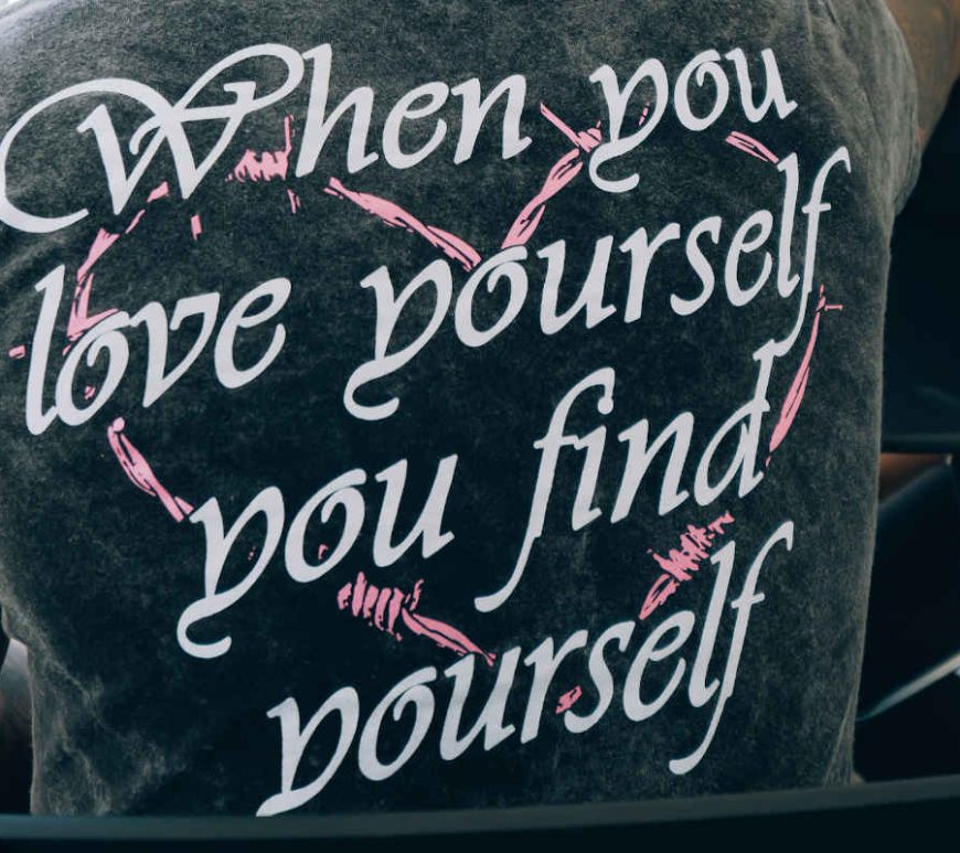 Back of a man's t-shift depicted. It is black stone-wash with a pink barbed wire heart and a quote in white overtop says "When you love yourself you find yourself".