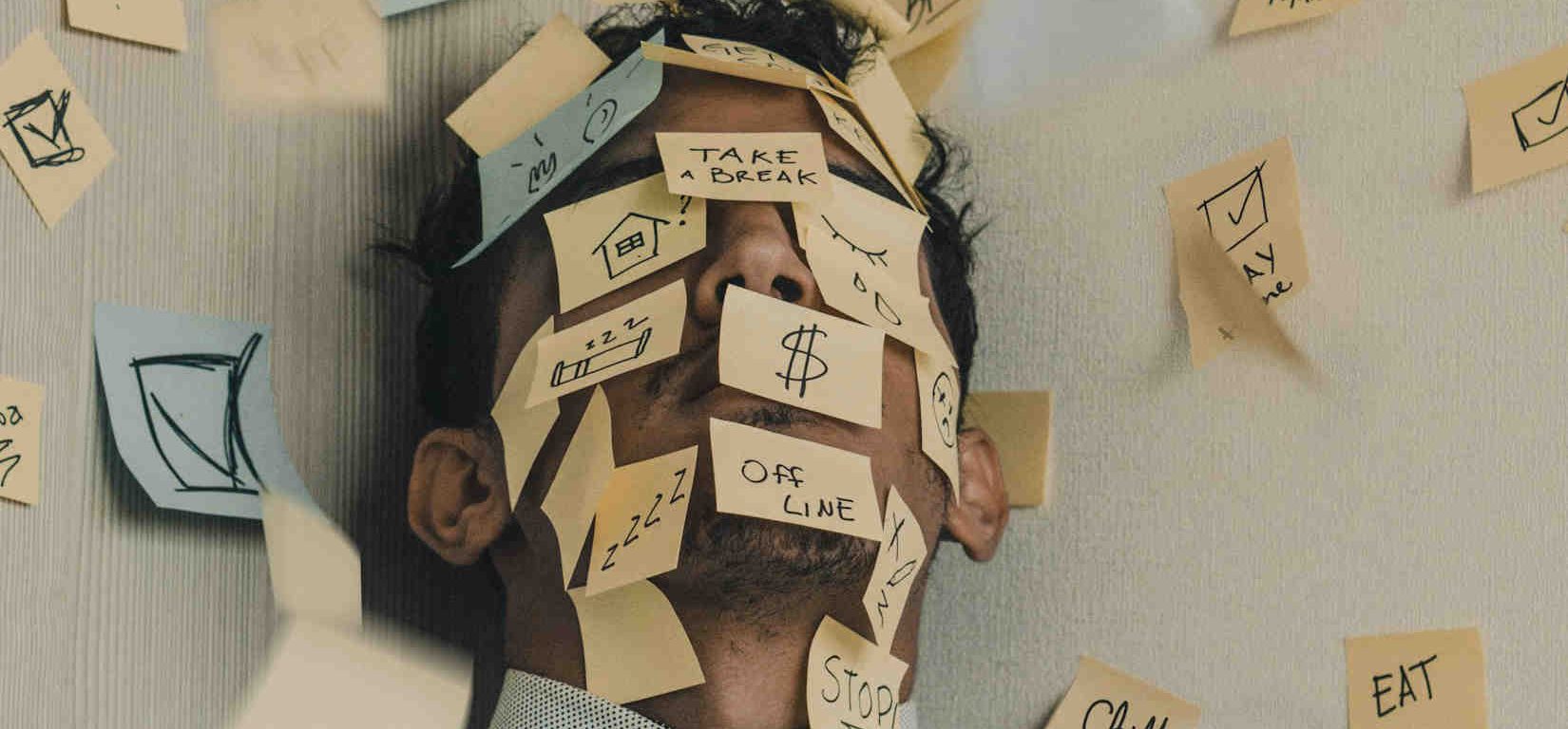 Man cornered between two walls with post-it notes all over his face and the walls. The notes have different responsibilities tied to work, home, finances, and self care.