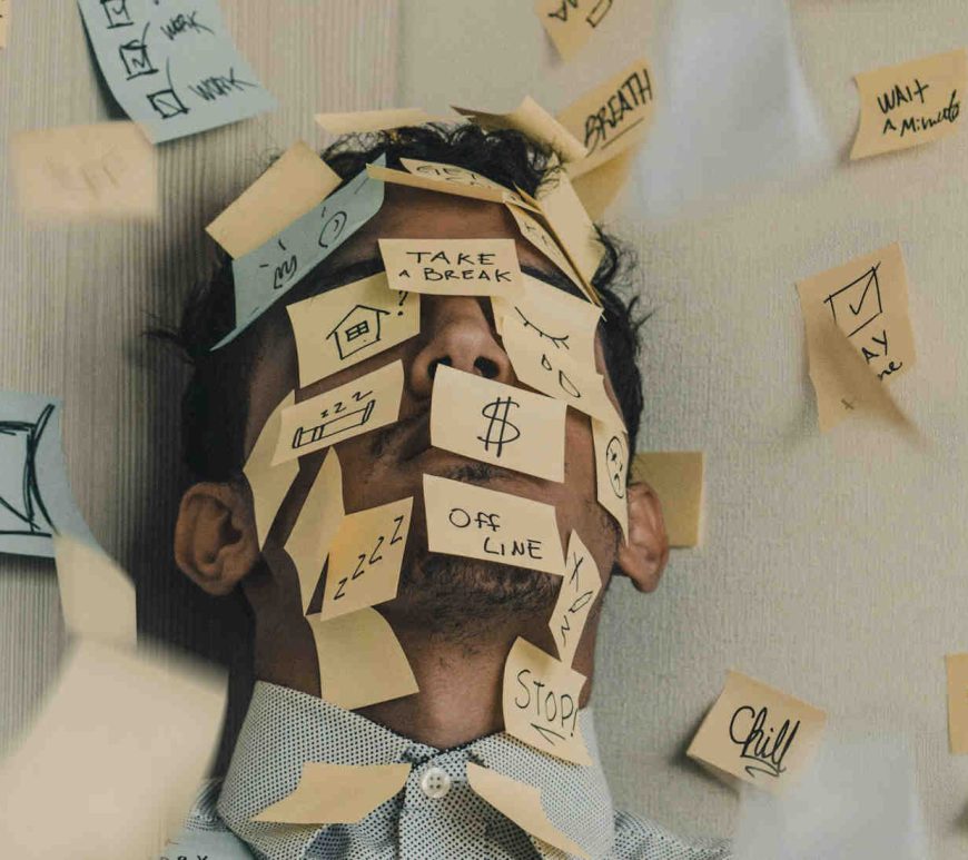 Man cornered between two walls with post-it notes all over his face and the walls. The notes have different responsibilities tied to work, home, finances, and self care.