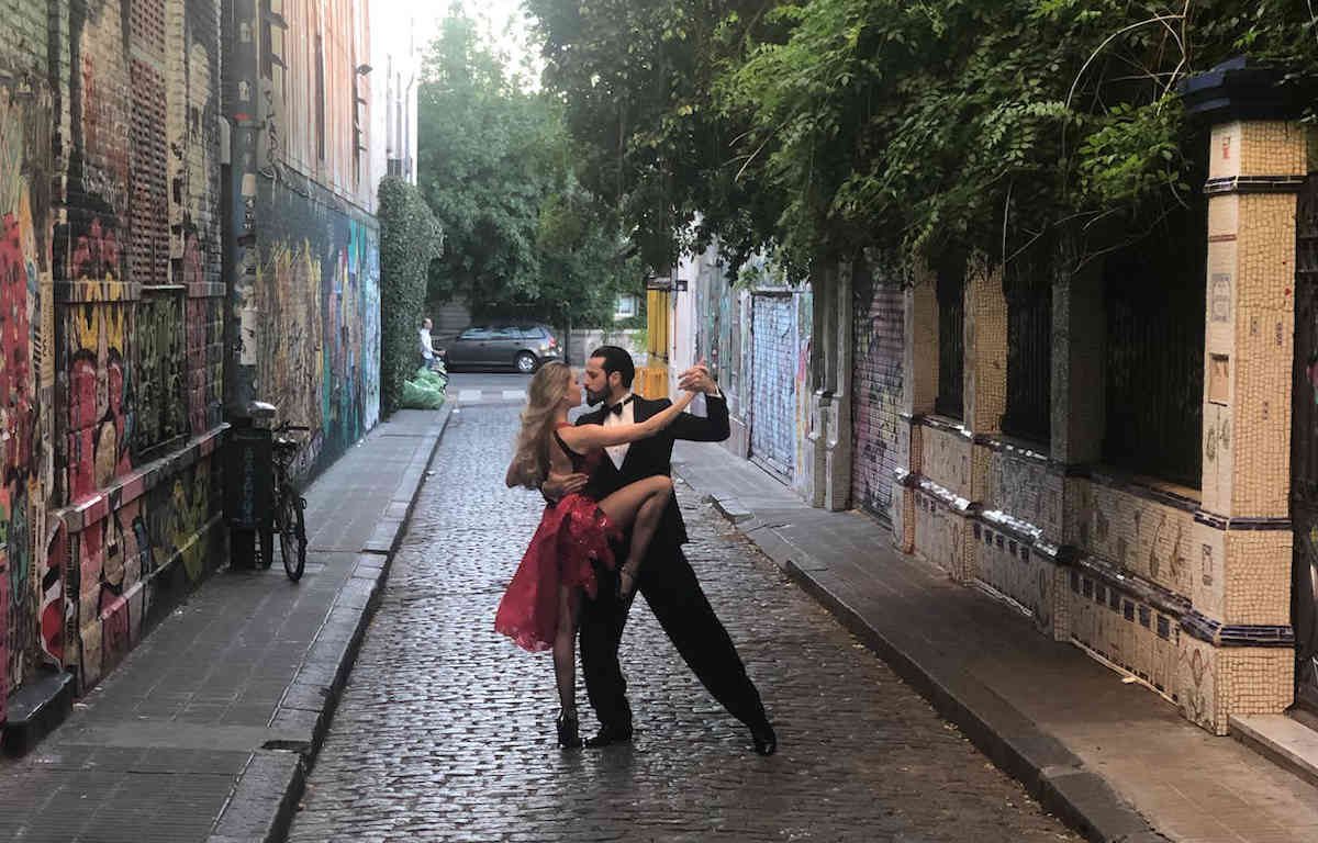 Man and woman dancing in an alleyway. The stone walls of the buildings are covered in graffiti and trees hang over them.
