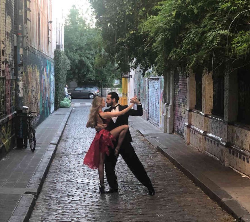 Man and woman dancing in an alleyway. The stone walls of the buildings are covered in graffiti and trees hang over them.