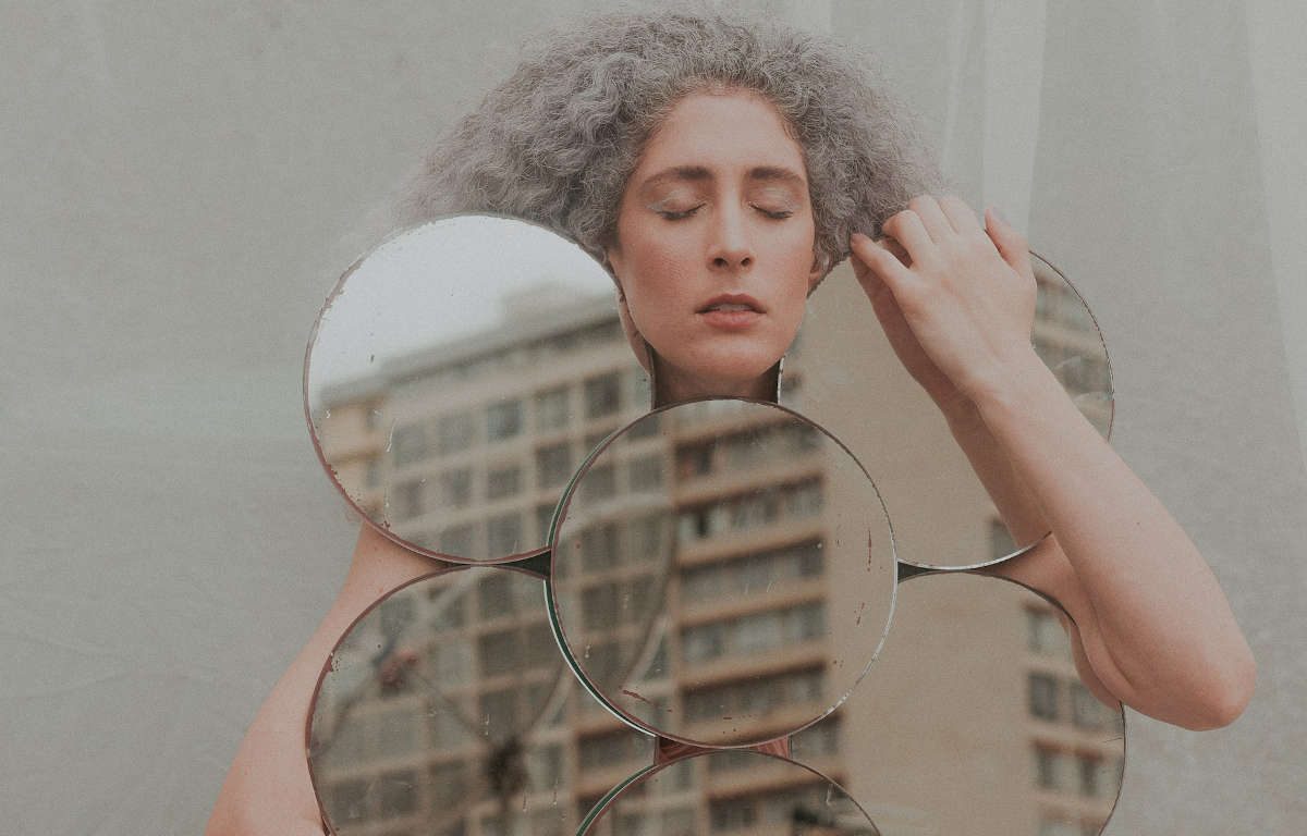 Photograph of a nude model with white, curly hair. Her eyes are closed and she is hugging an artistic mirror of circles.