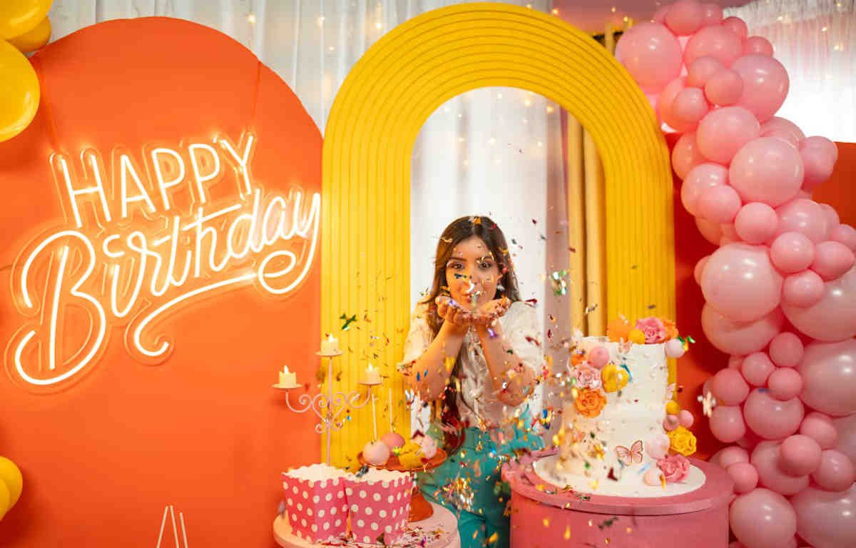 Woman blowing confetti under a yellow arch in a room with pink balloons, a celebratory cake, and a neon Happy Birthday sign against an orange backdrop.