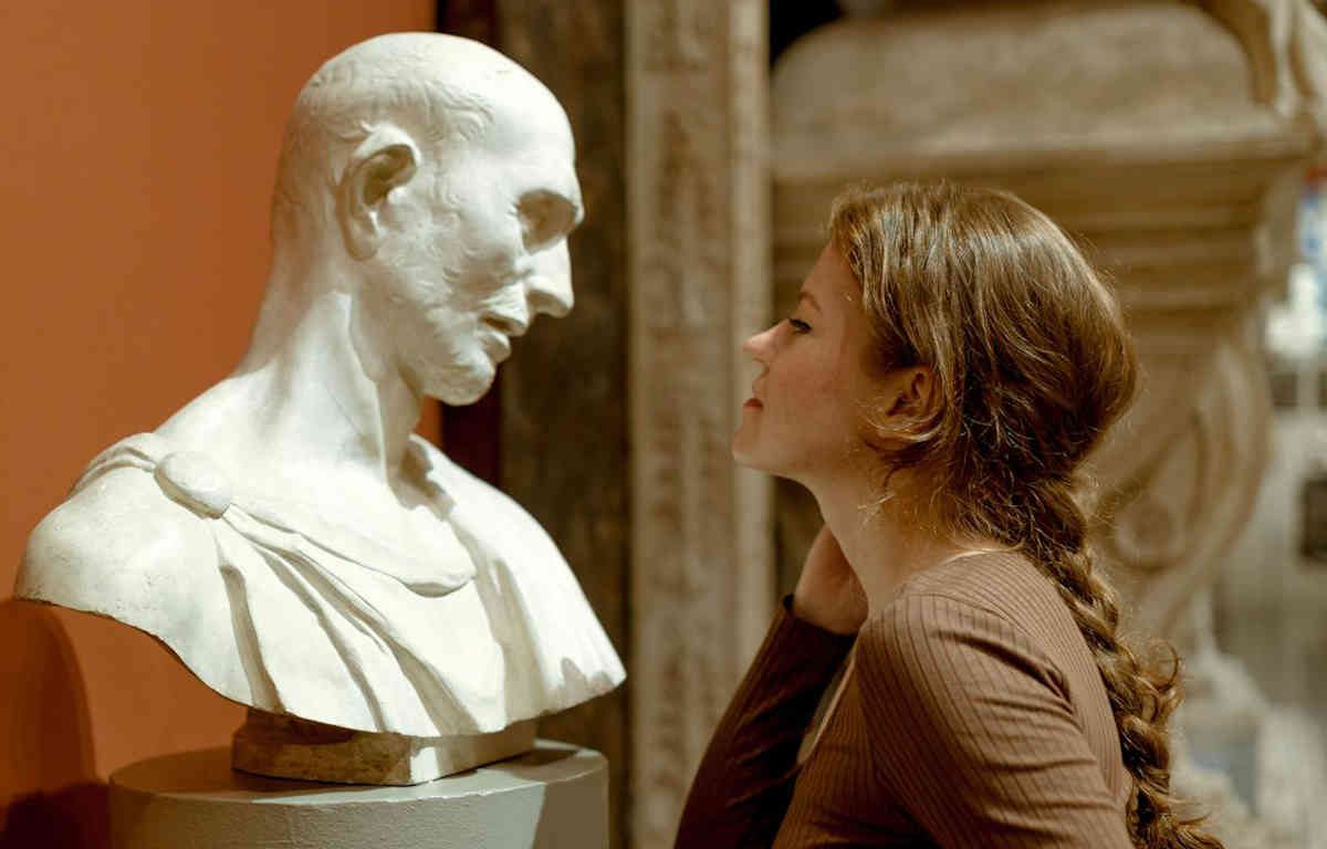 Woman looking at bust of man on pedetal with admiration.
