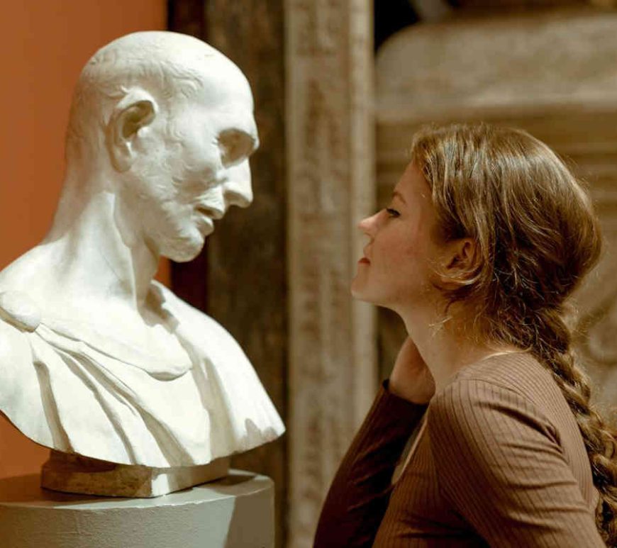 Woman looking at bust of man on pedetal with admiration.