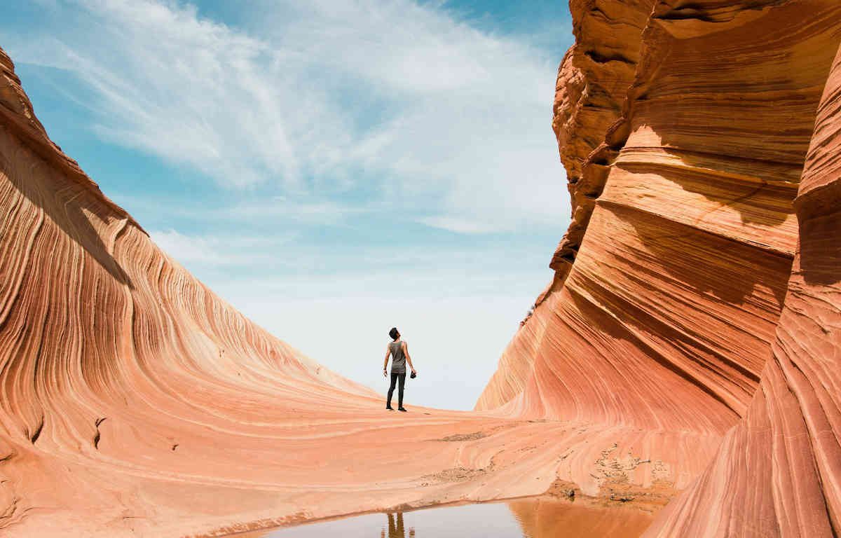 Man standing at base of Antelope Canyon canal staring up in awe. The canyon is red, the sky in the background is blue with dispersed clouds.