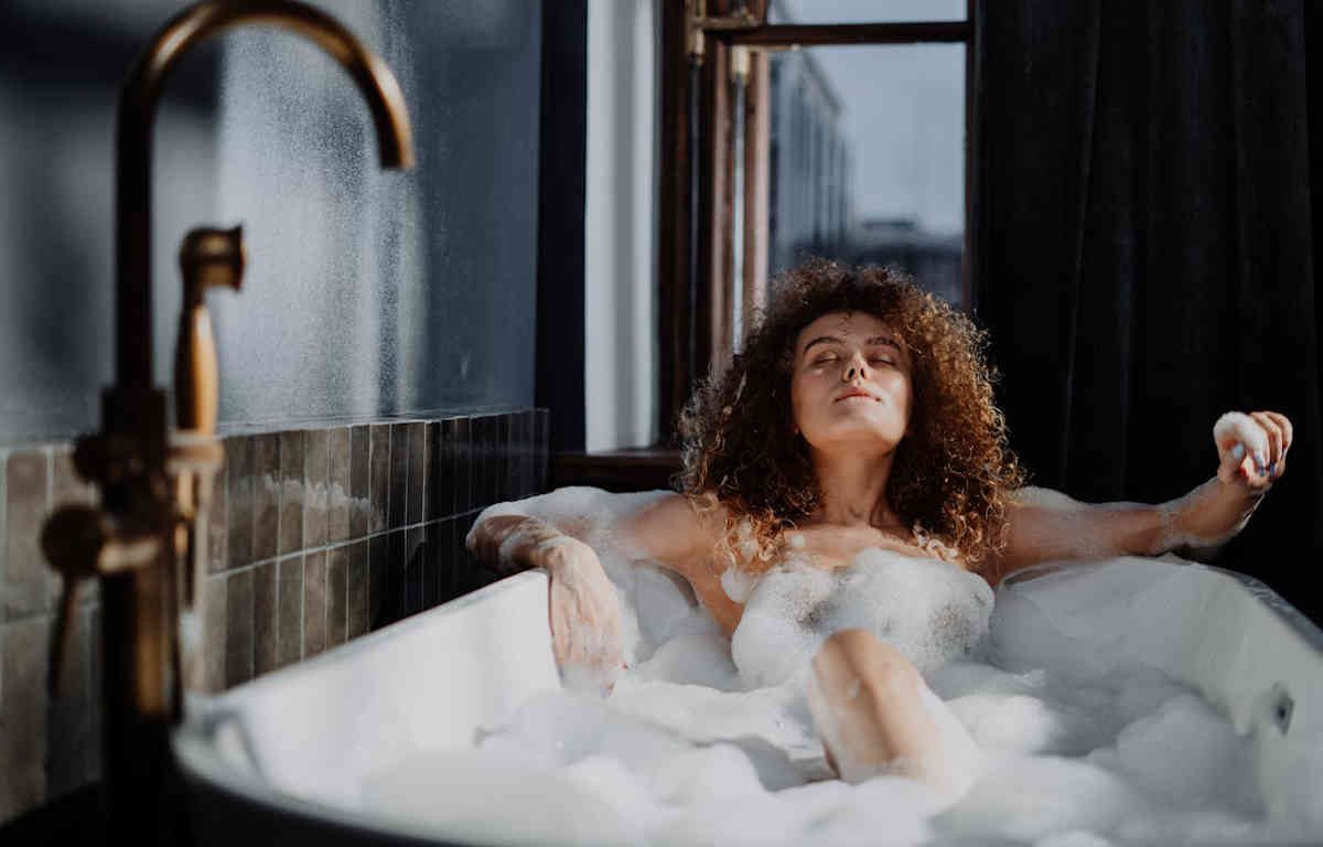 Woman soaking in a bubble bath. Her skin glows with the gentle sunlight coming through a window.