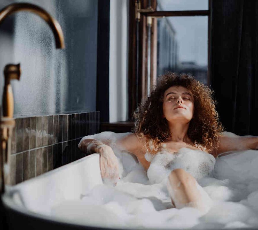 Woman soaking in a bubble bath. Her skin glows with the gentle sunlight coming through a window.