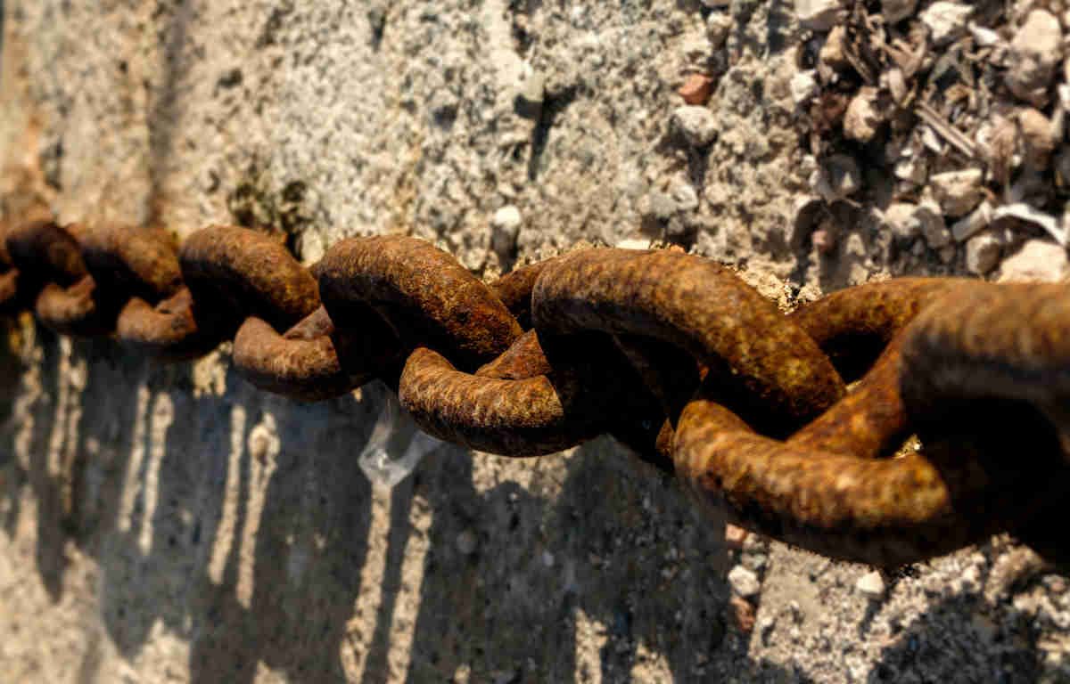 Rusted chain links. The sun casts a shadow on the rocky ground beneath.