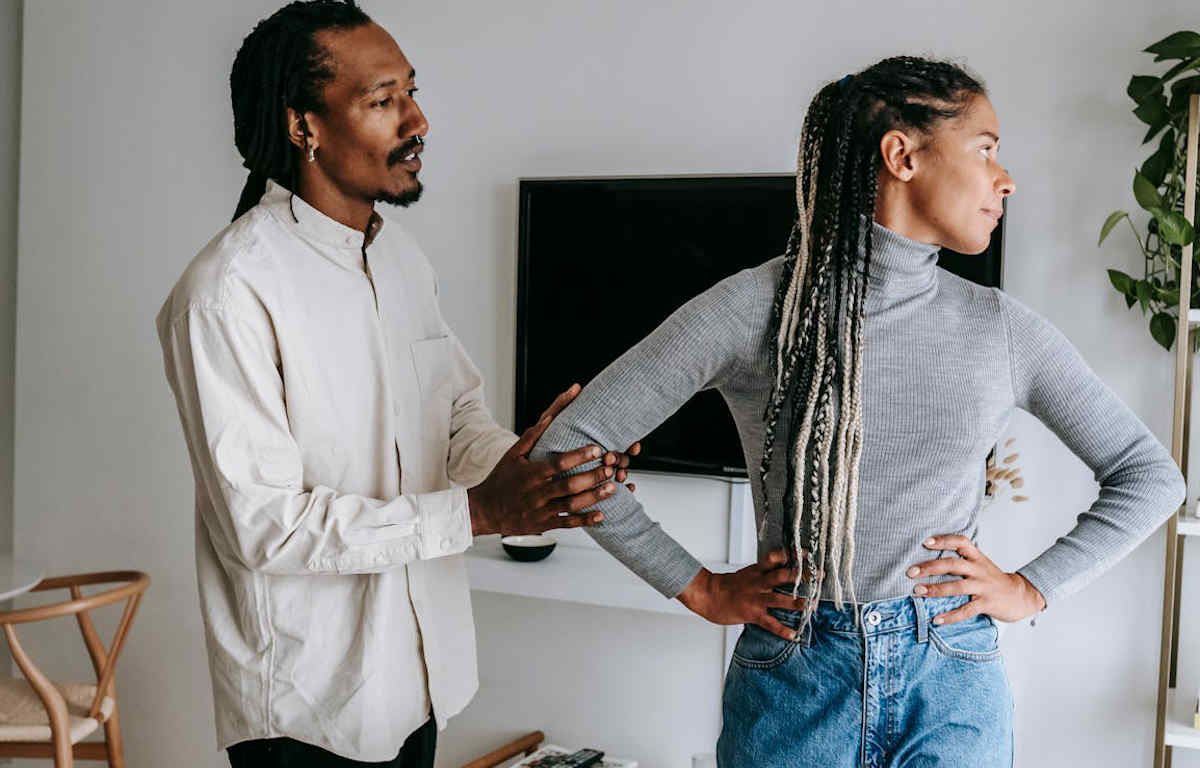 Black man trying to talk to a black woman that is facing away from him upset. Her arms are at her waist, he is holding her right arm with his hands.