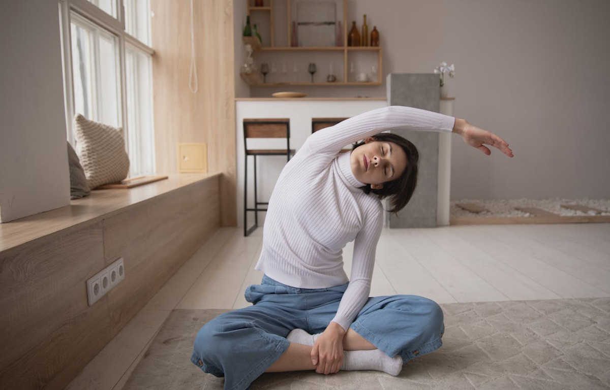 Woman sitting on the floor with legs crossed, stretching her arm over her head to the opposite side. Her eyes are closed. The room has natural light and soft tones creating a relaxed background.