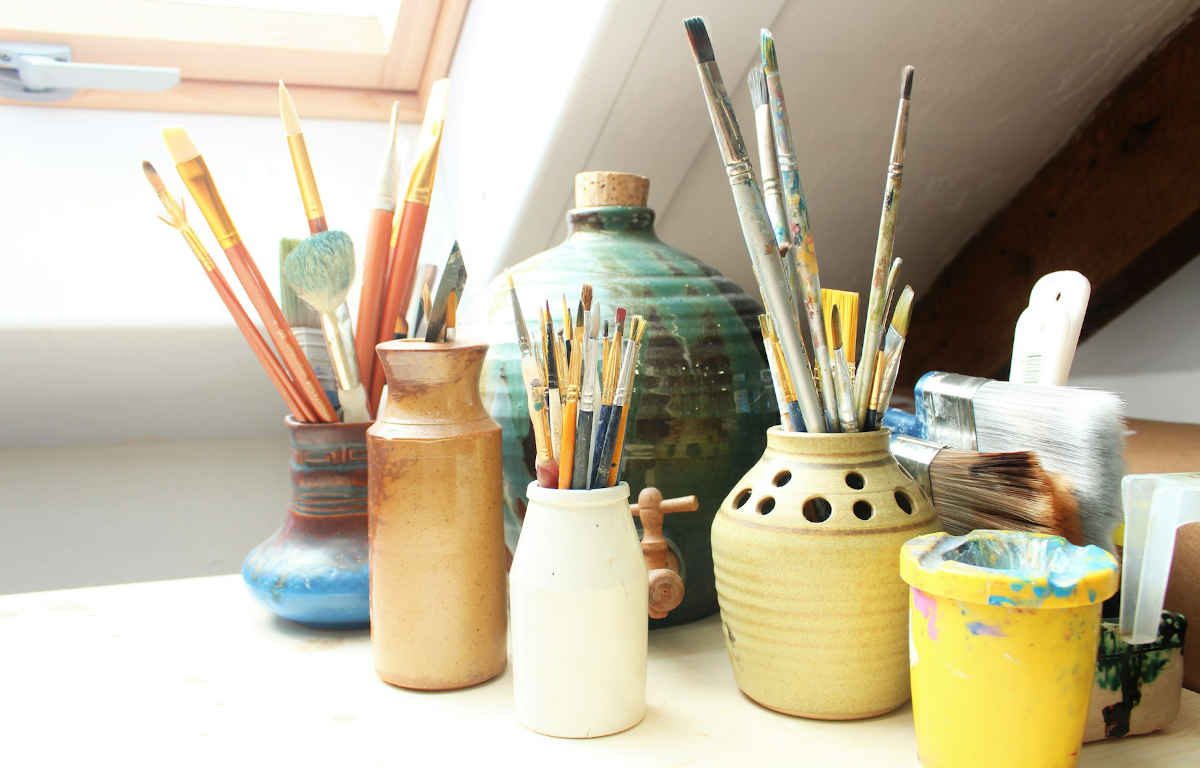 Tabletop with assorted paint brushes in handmade ceramic vessels. Natural light is coming through the window.