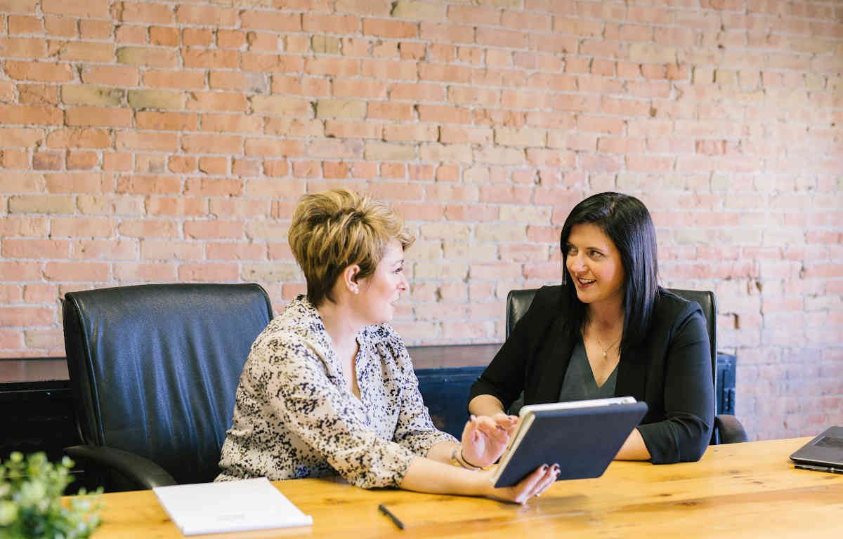 Two women having a meeting. They are sitting at a large wood table with black leather board room chairs.