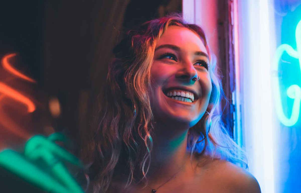 Young lady with big smile looks onto the distance. Her skin glows with the reflection of neon lights.