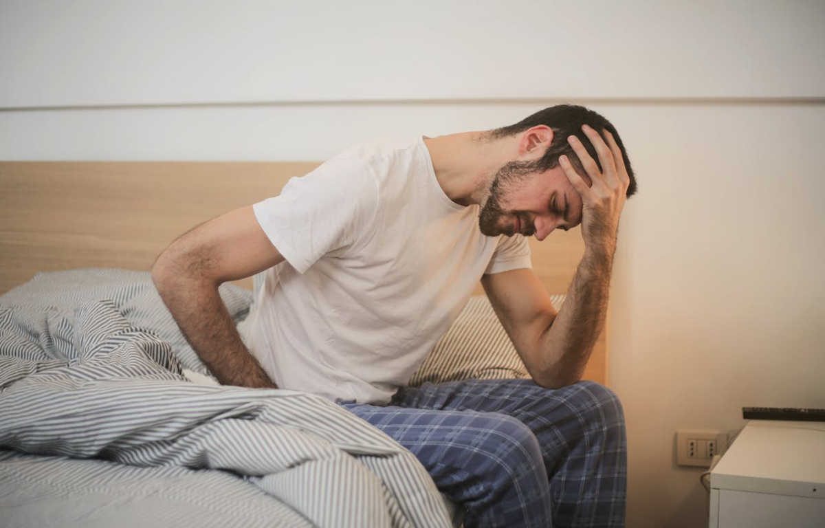 Man in pyjamas sitting on the edge of the bed holding his side and his head.