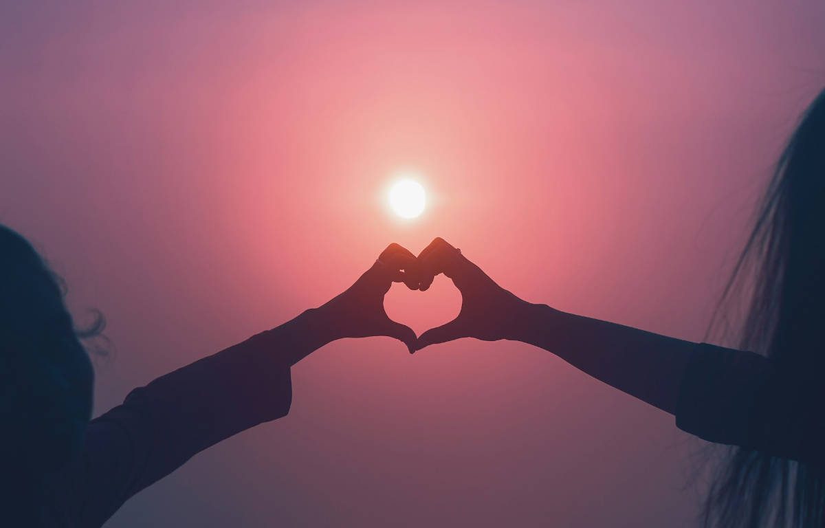 Shadow of two people forming a heart shape by putting their hands together with a sunset in the background.