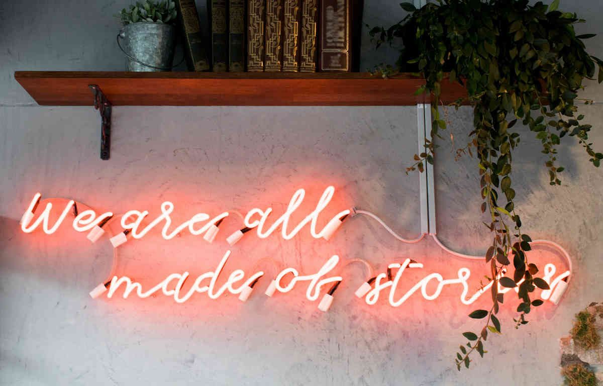 Neon text on a wall below a wood shelf with books and plants reads "We are all made of stories".