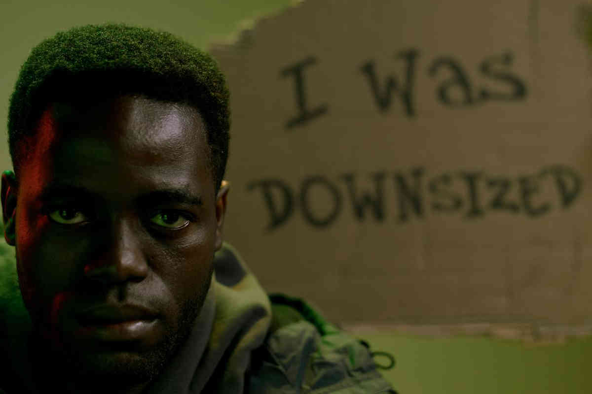 Black man starring forward with sadness in his eyes and a cardboard sign behind him saying "I was downsized".