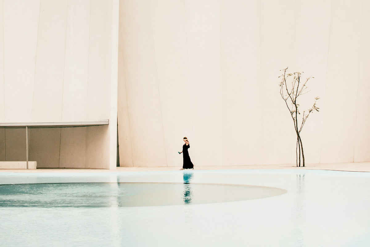 Woman in long black dress walking by outdoor water pool next to a massive white wall with a single, woody tree next to it.